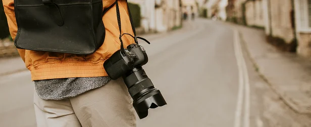 Een persoon in een oranje jas en met een rugzak houdt een camera vast op een rustige, lege straat omringd door pittoreske huizen, wat een gevoel van verkenning oproept.