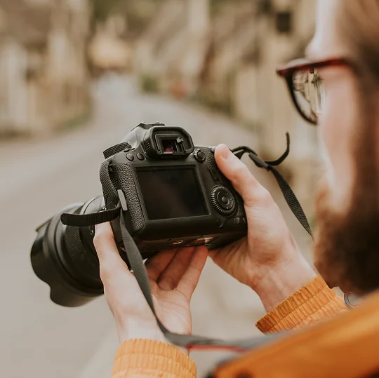 Een persoon met lang haar, die een bril draagt en een oranje jas aan heeft, houdt een DSLR-camera vast op een vervaagde straat, wat de focus op fotografie suggereert.